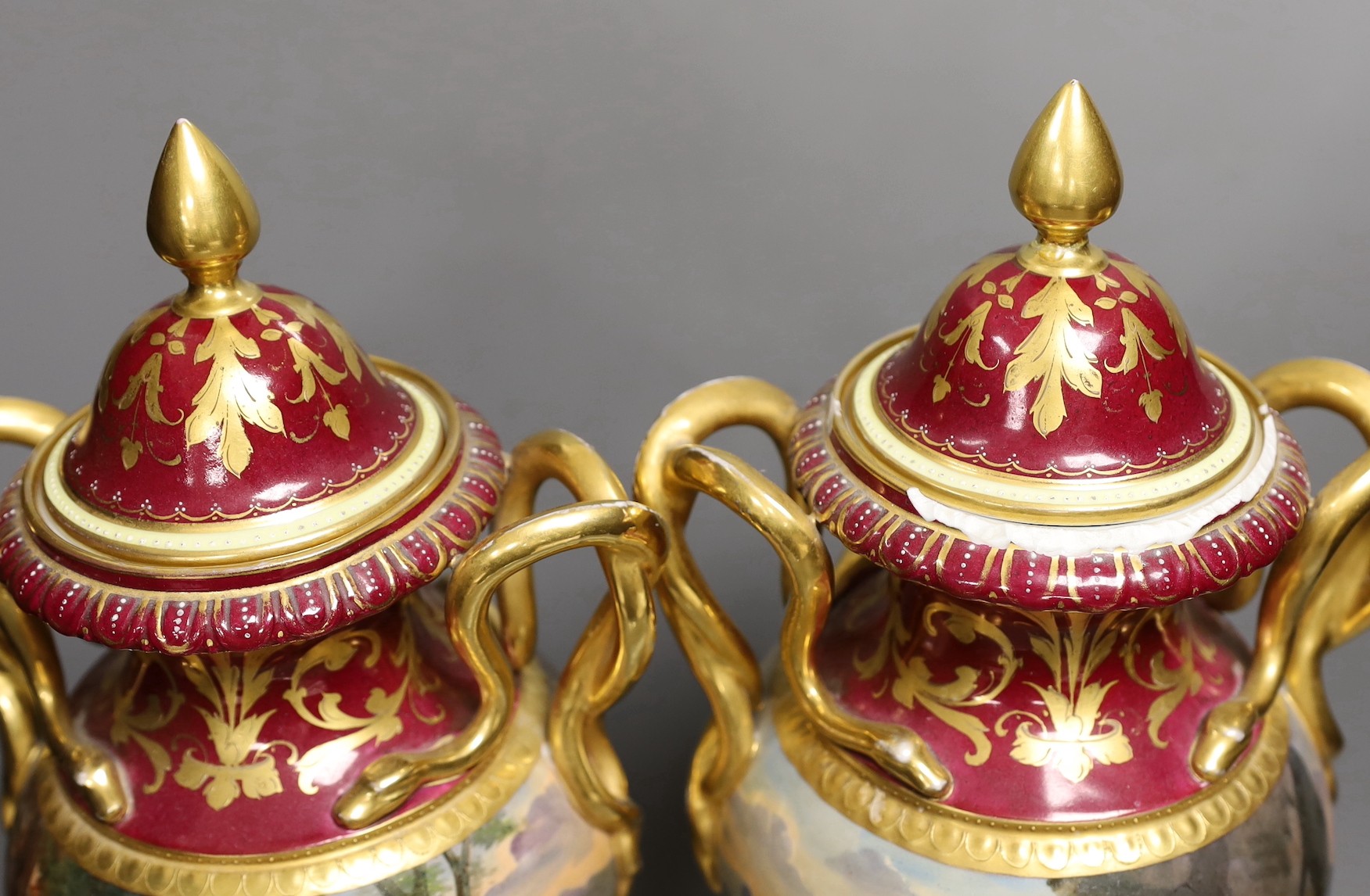 A pair of ornate Vienna style two handled urns bearing covers, with painted love scene and red ground and gilt decoration, 46.5cm high